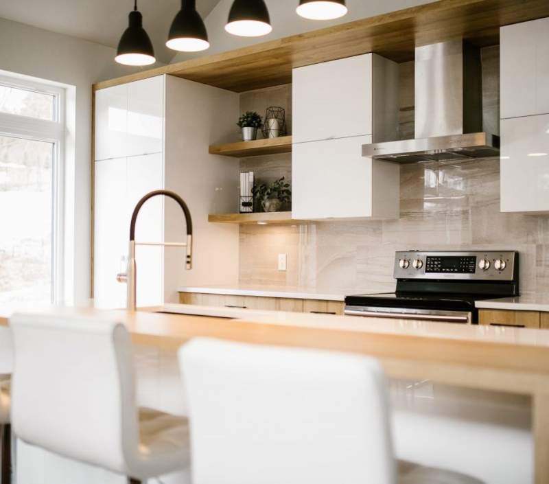 This modern white kitchen has wood accents, two chairs, appliances, potted plants, and decorated countertops for a complete, cozy aesthetic.